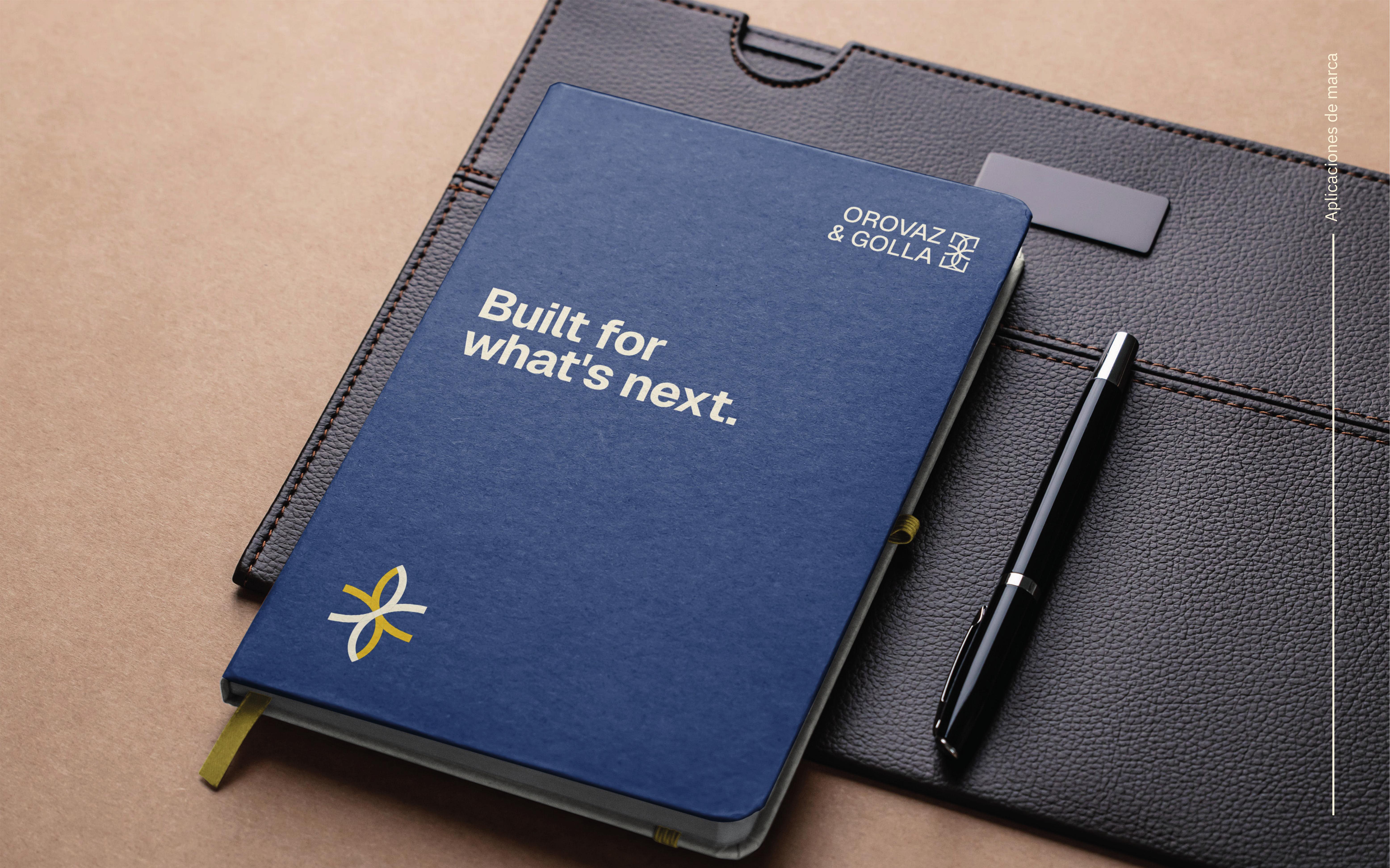Cuaderno azul con texto 'Built for what’s next' junto a un bolígrafo negro sobre una superficie marrón.