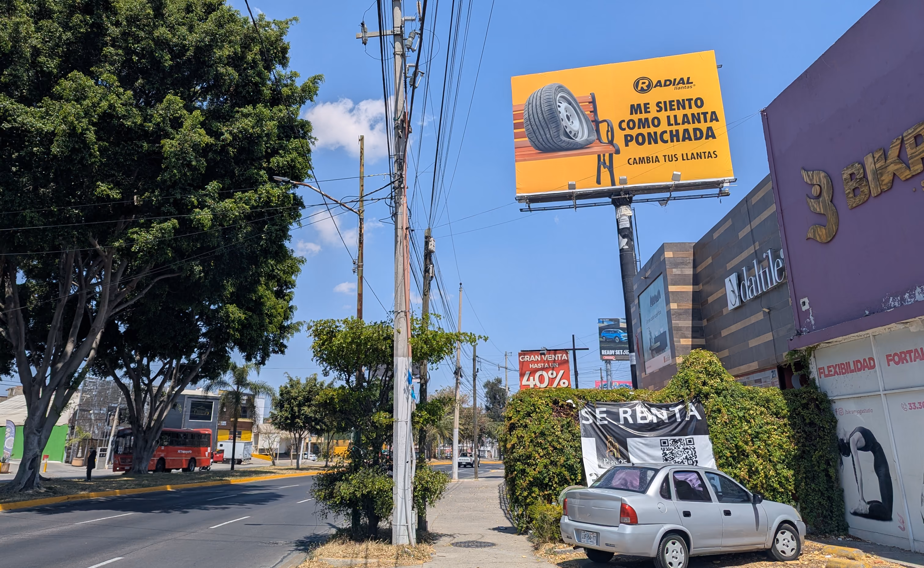 Calle con árbol grande, coche gris estacionado, y anuncios de venta y cambio de llantas en México.