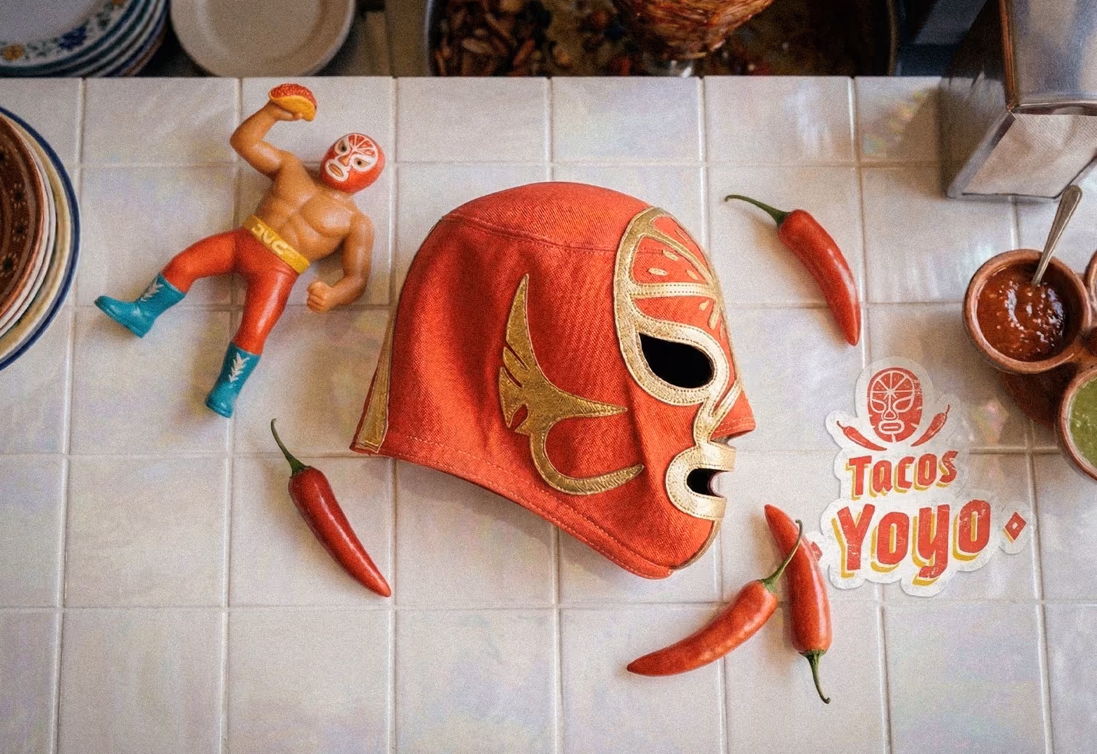 Máscara roja de luchador y figura con máscara sosteniendo taco sobre mesa con chiles y salsas.