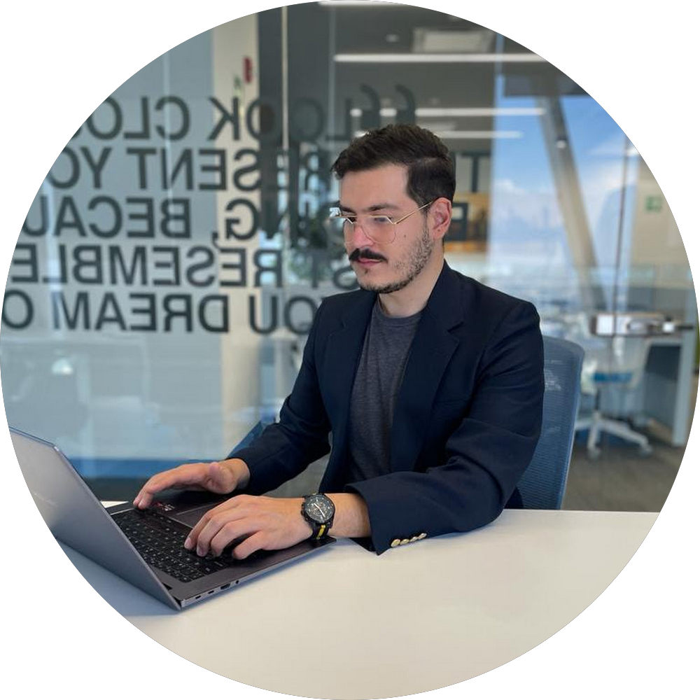 Hombre con gafas y saco trabajando en laptop en oficina moderna con paredes de vidrio.