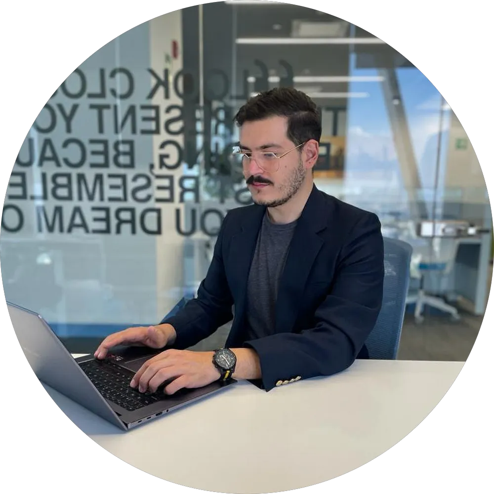 Hombre con gafas y saco trabajando en laptop en oficina moderna con paredes de vidrio.